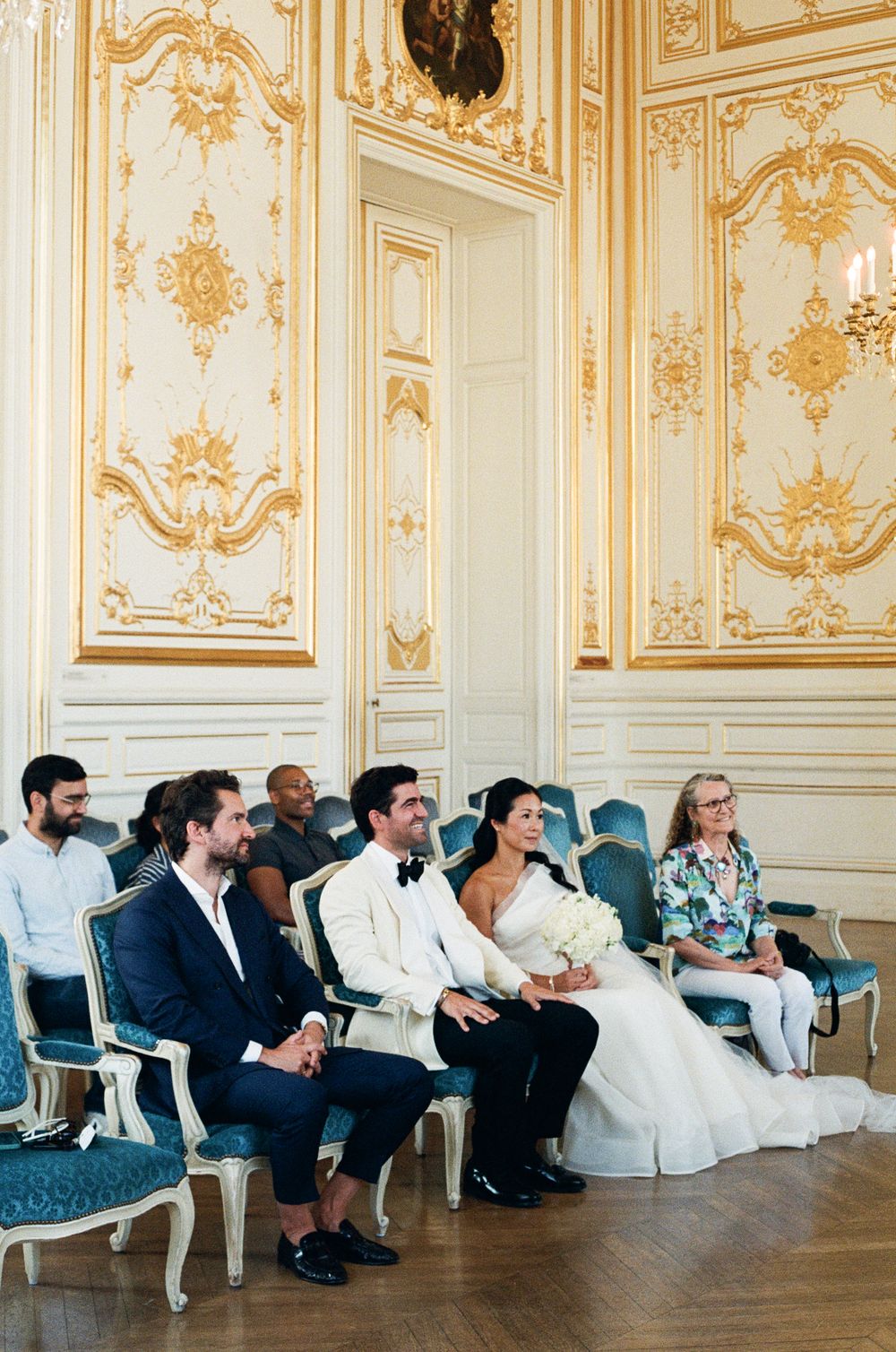 Bride and groom seated at elegant French wedding ceremony in ornate gold and white ballroom with blue velvet chairs.