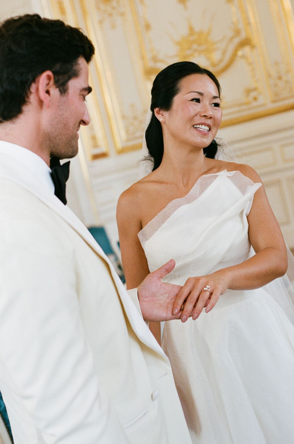 Bride in white one-shoulder gown smiles at groom in white tuxedo during Versailles wedding. Versailles ceremony hall, wedding phtoographer.