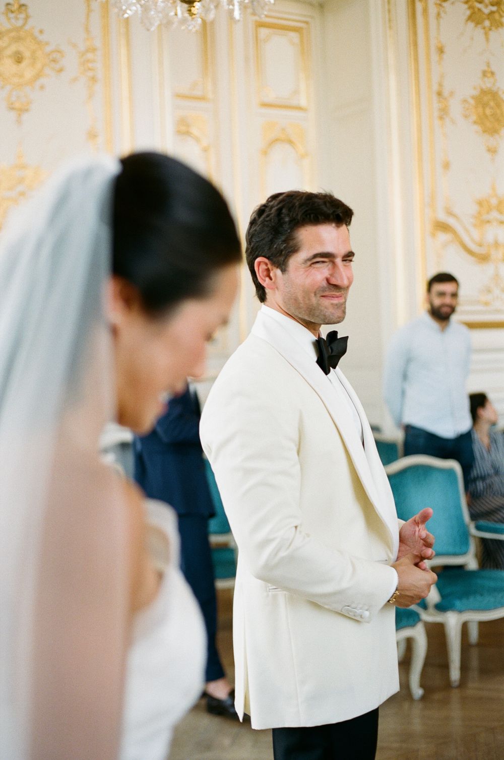 Groom in white tuxedo smiling at bride during elegant wedding ceremony in ornate gold ballroom. Versailles mairie, Versailles wedding