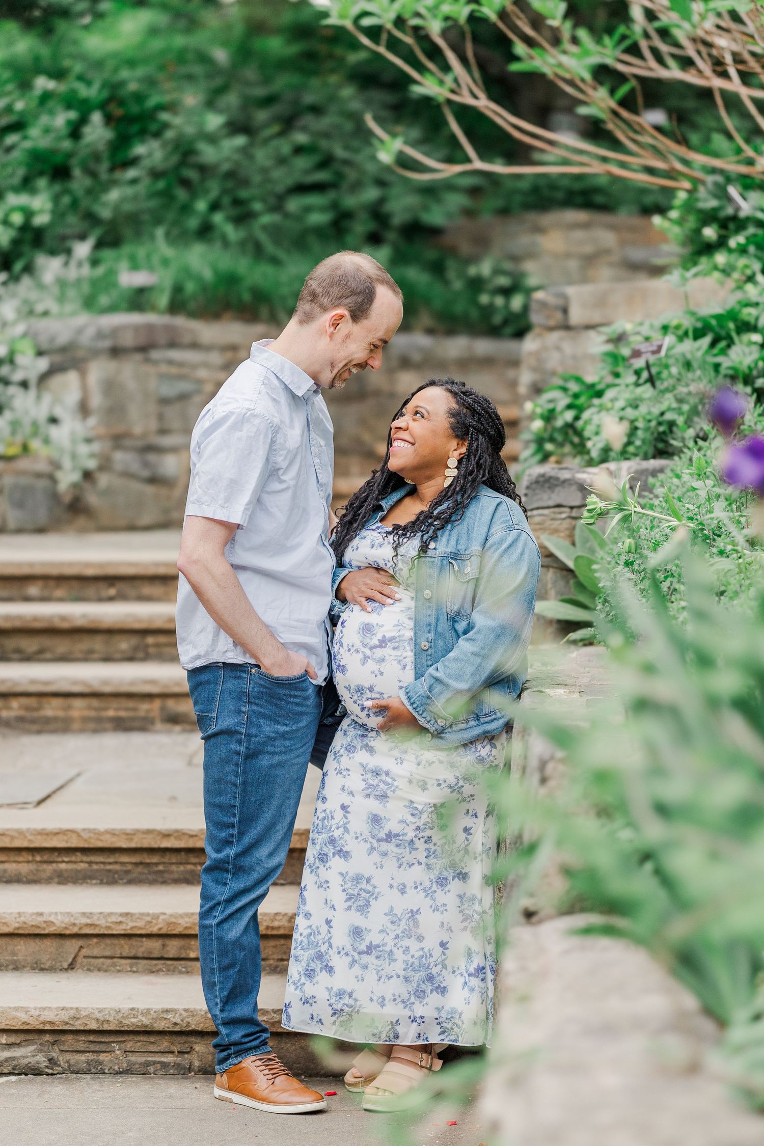 Pregnant woman in floral dress and denim jacket smiles at partner on garden steps surrounded by lush greenery.