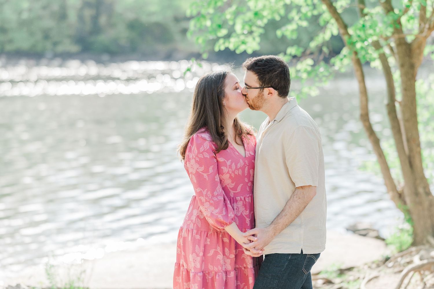 Couple sharing a romantic kiss by a sunlit river, woman in pink floral dress, man in beige shirt, surrounded by green trees.