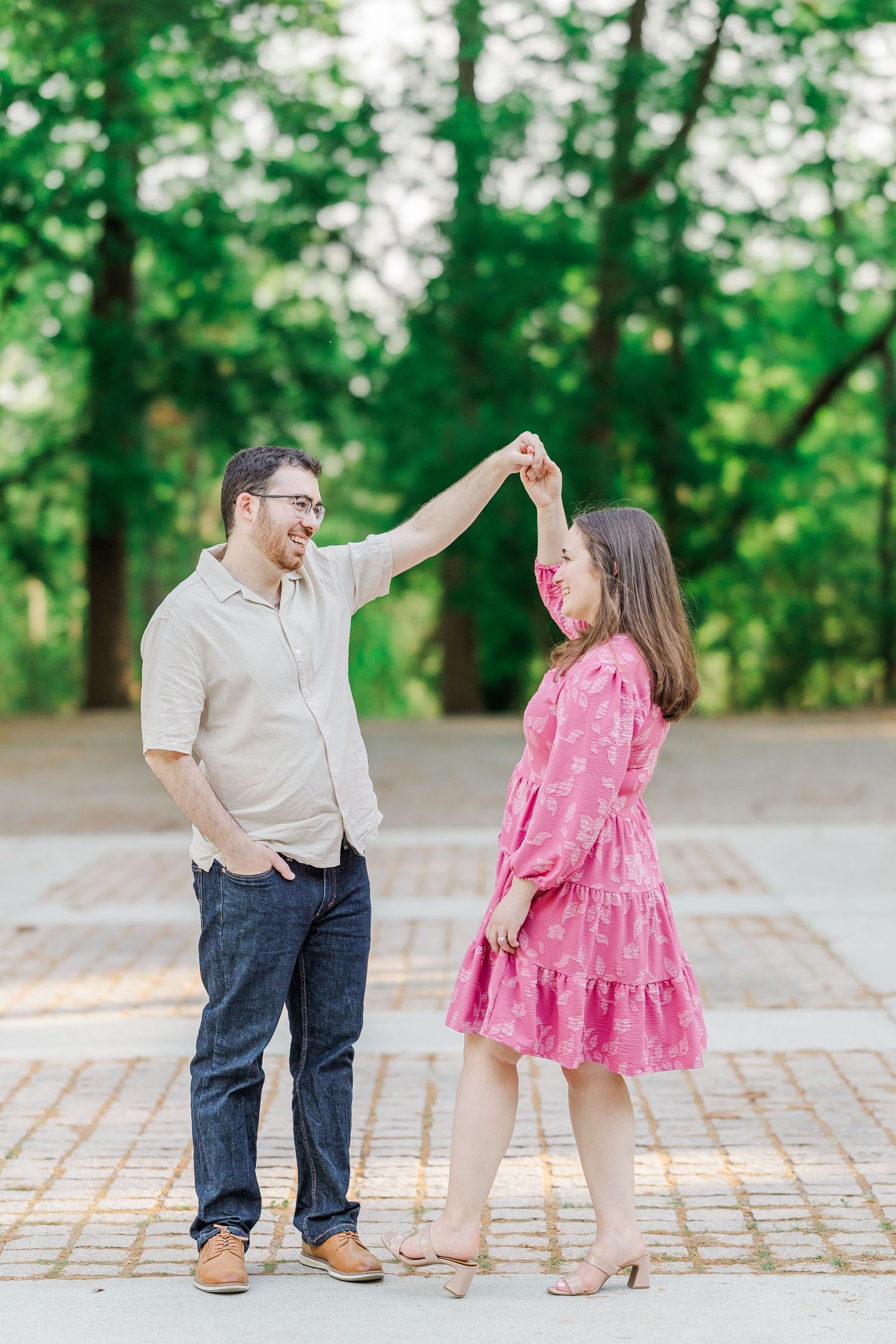 A couple dances outdoors in a park, man in beige shirt and jeans twirling woman in pink floral dress.