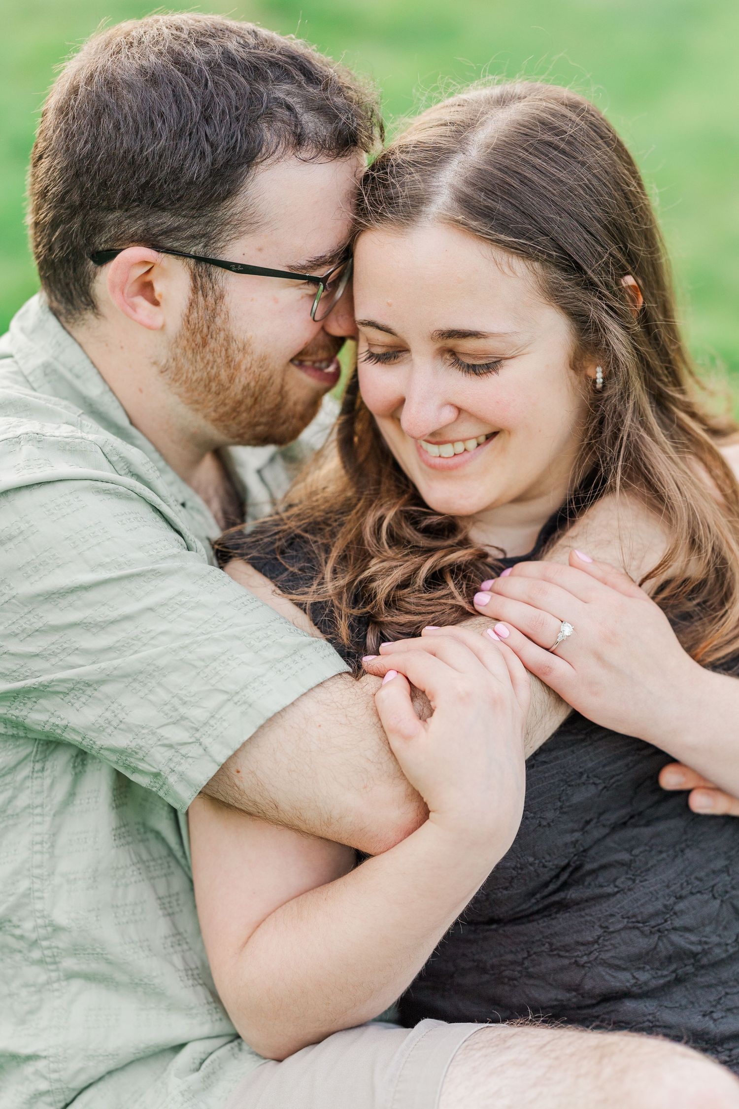 A couple shares a tender embrace outdoors, the woman smiling as the man nuzzles close, her engagement ring visible.