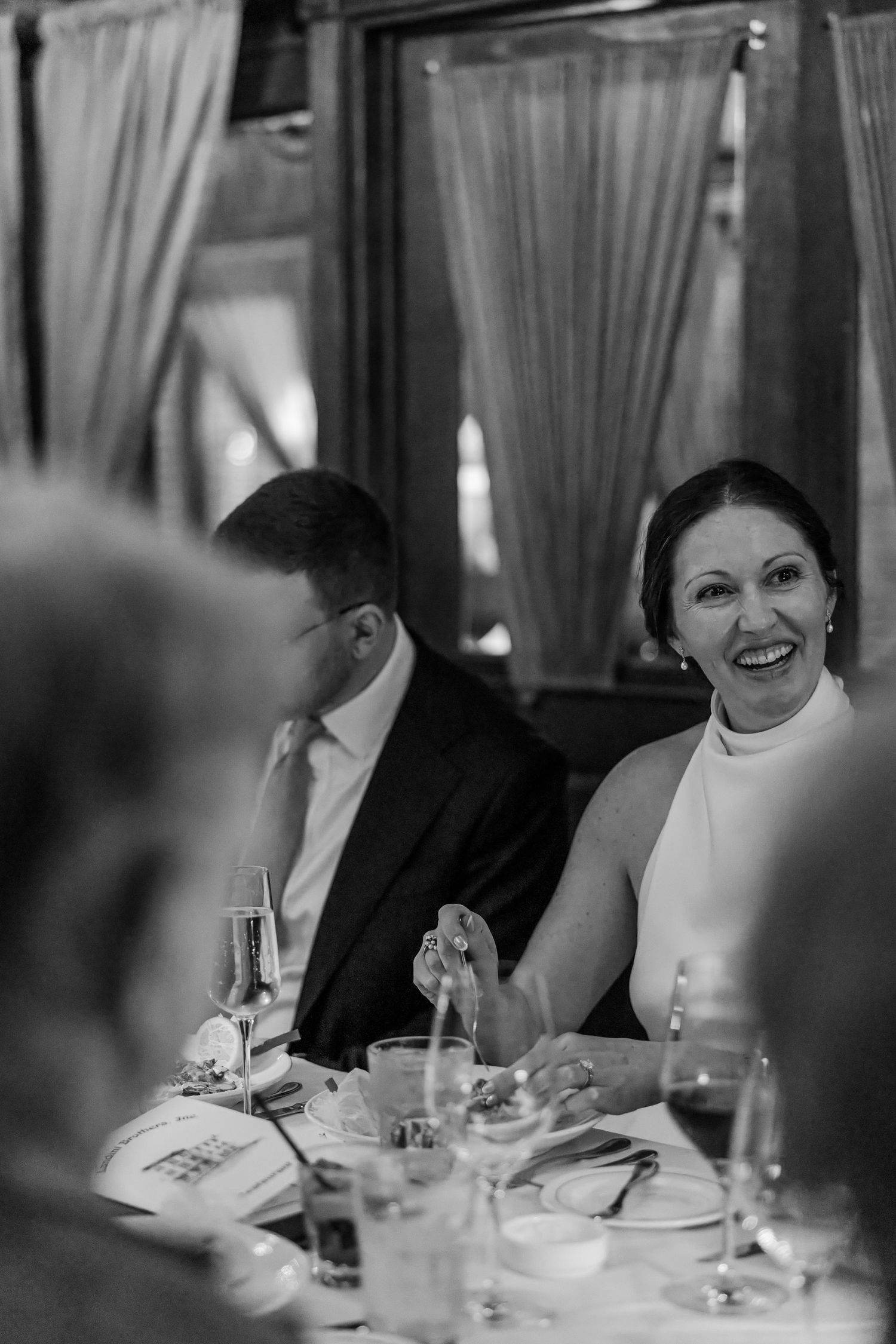 Black and white photo of a smiling woman in a white dress at an elegant dinner table with champagne glasses.