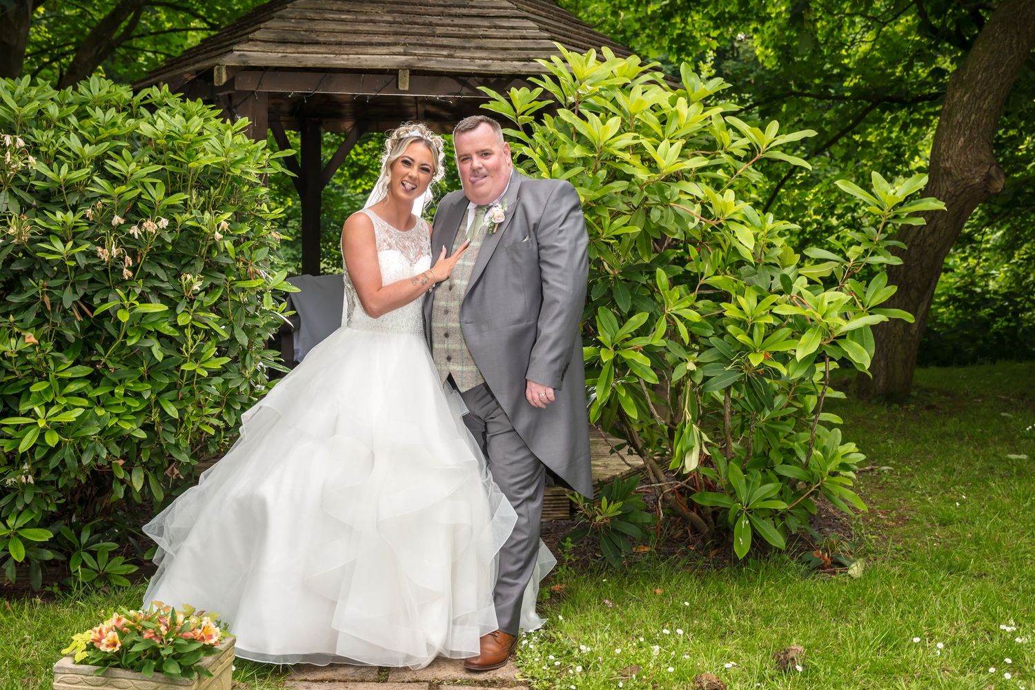 Bride in white gown and groom in grey suit pose together in a lush garden setting on their wedding day.