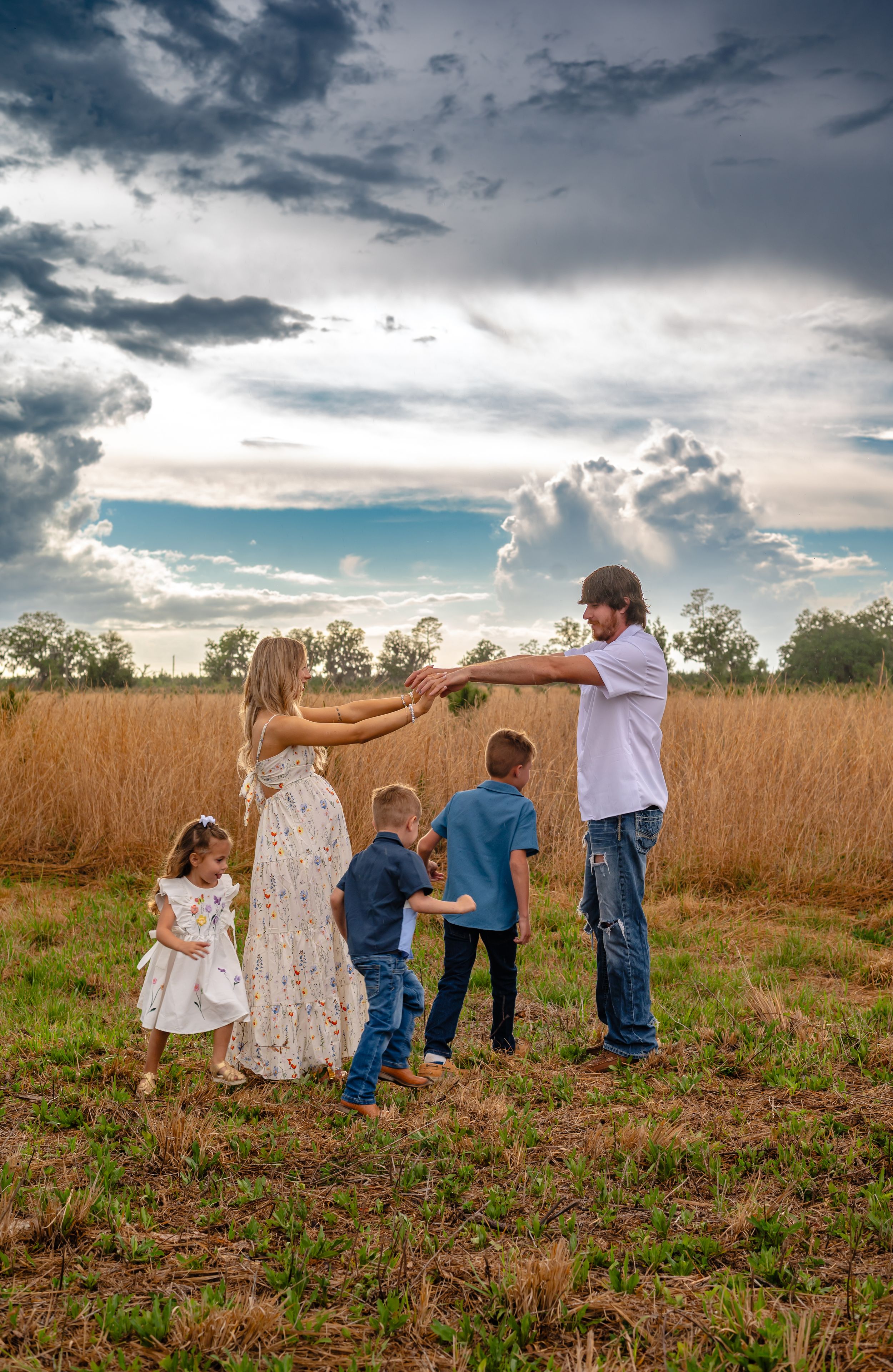 Family of five playing in a golden wheat field under dramatic storm clouds at sunset in a rural countryside setting.