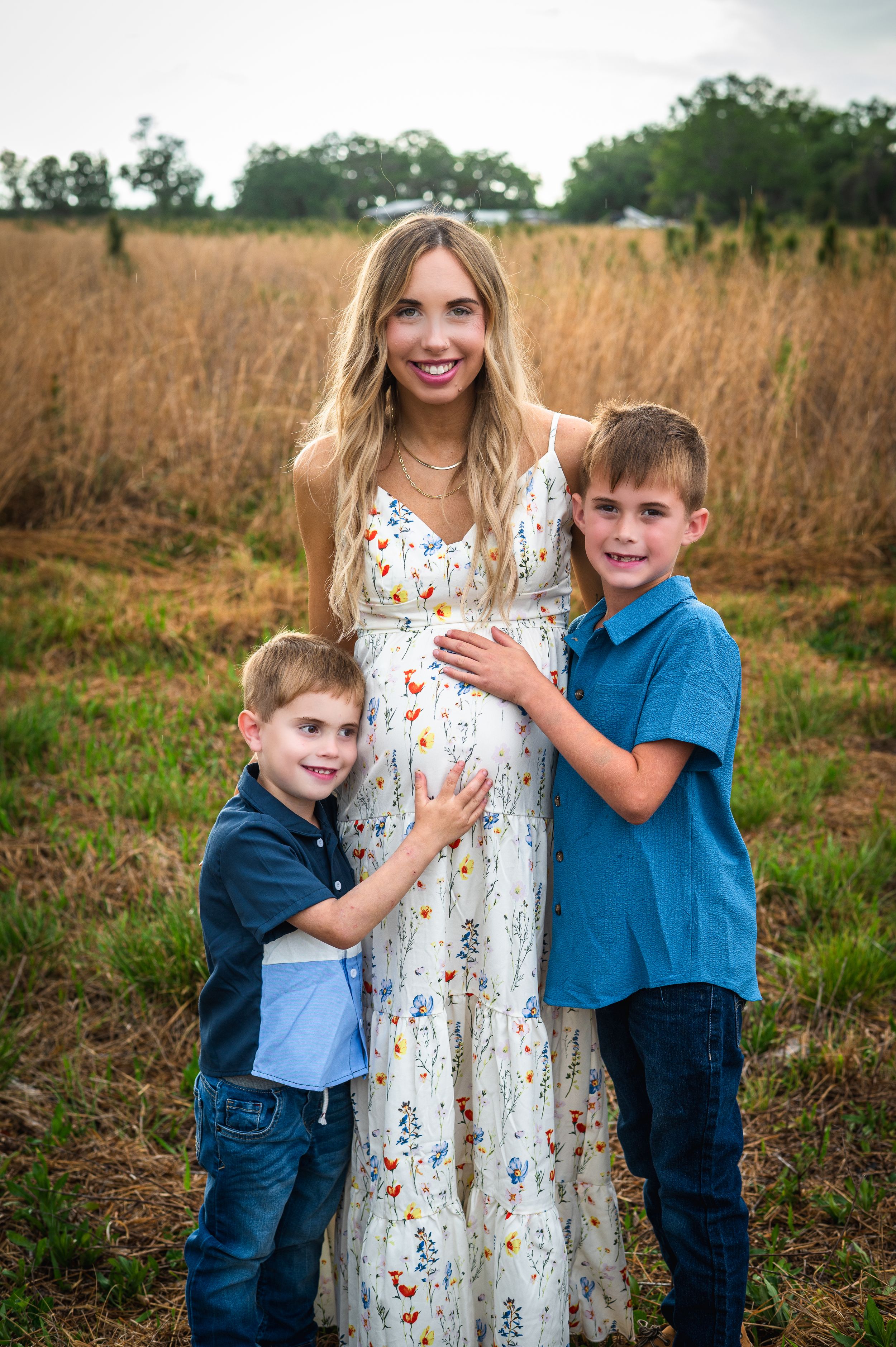 Pregnant woman in floral maxi dress poses outdoors with two young boys hugging her baby bump in a golden field.