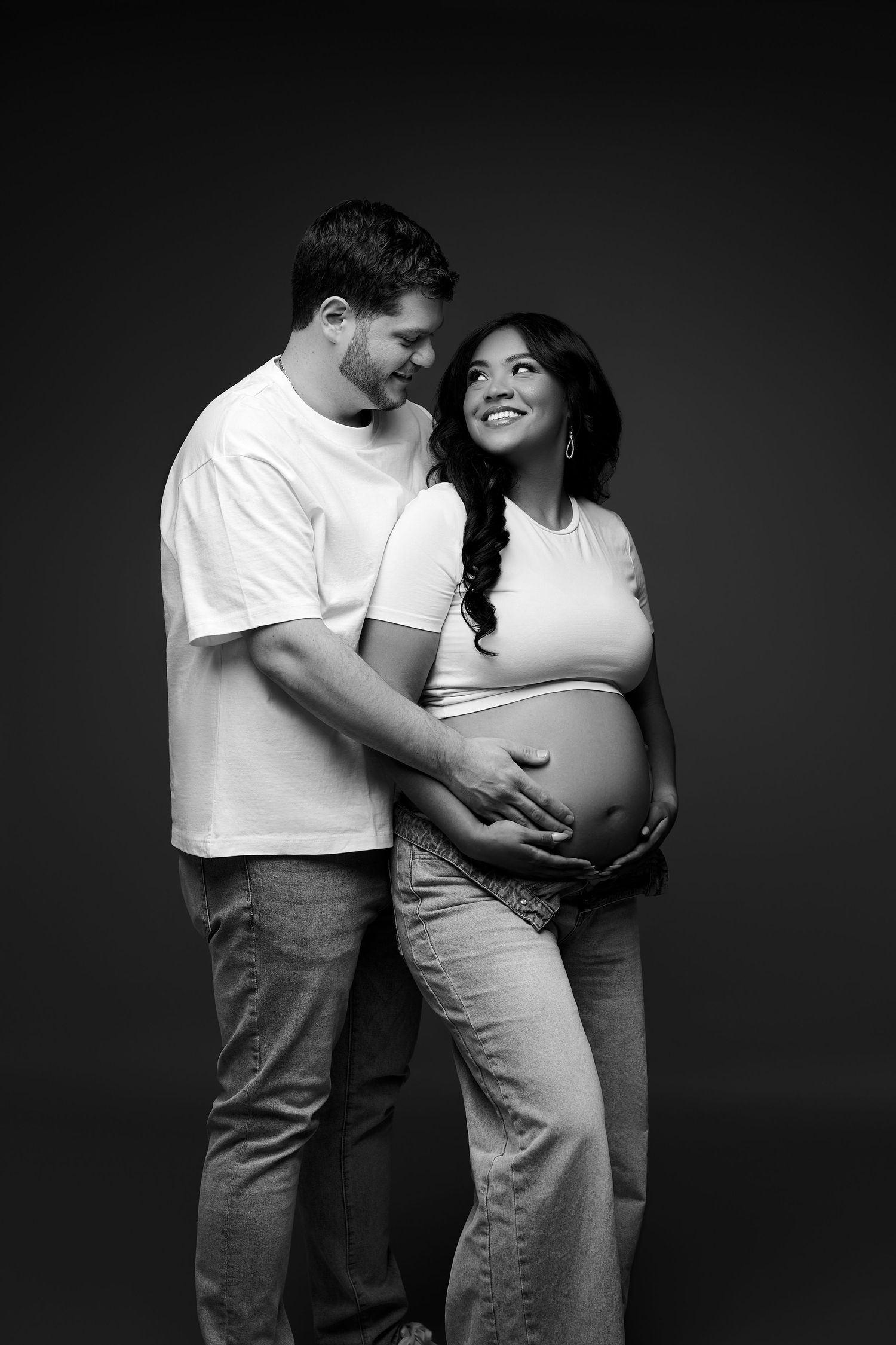 Black and white maternity photo of a smiling couple in white shirts cradling pregnant belly in studio.