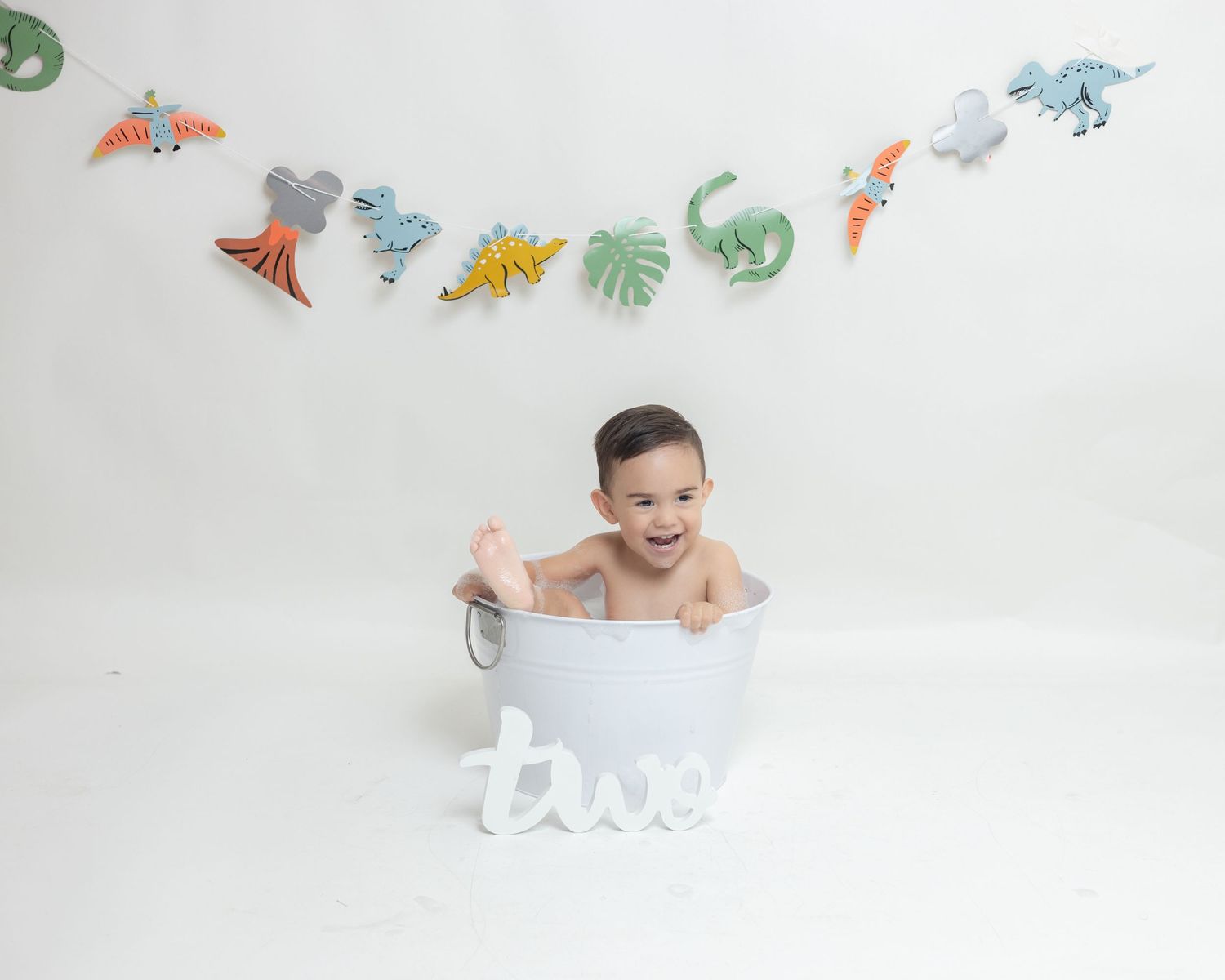 Smiling baby sitting in a white tub with dinosaur banner and 'two' letters for a second birthday photo.