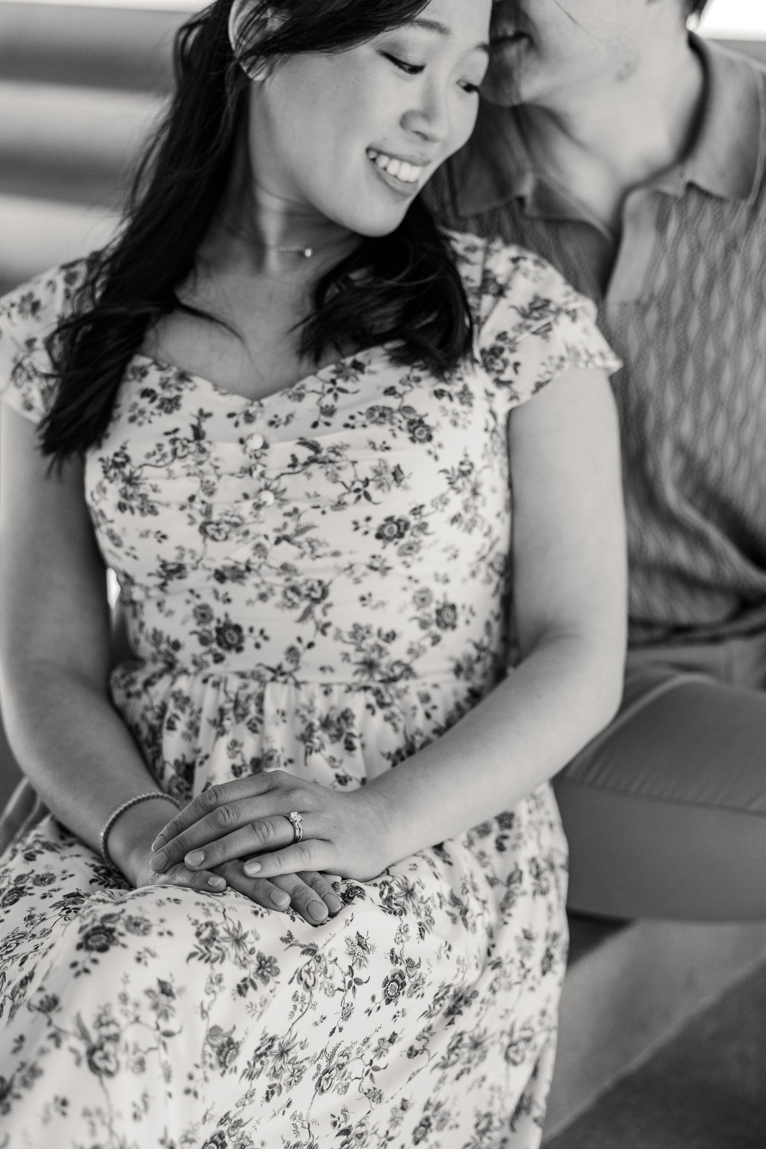 Black and white photo of a smiling woman in floral dress sitting closely with a man, showing an engagement ring.