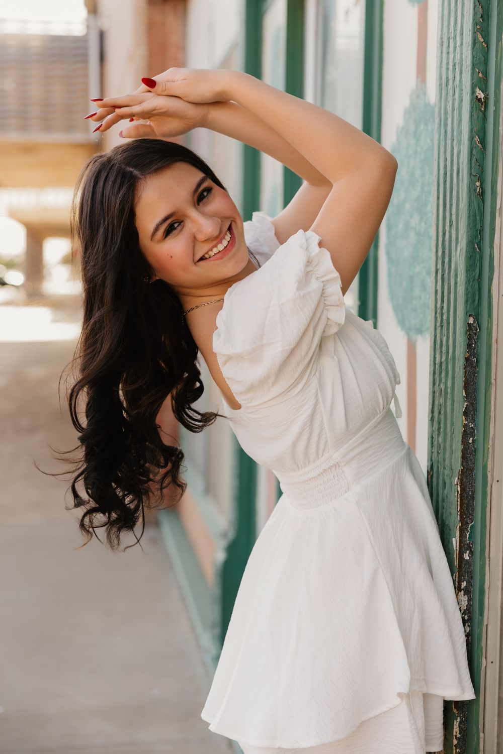 Brunette girl wearing white dress smiles for photos in downtown Tulia, Texas and stretches her arms over her head for a fun pose.