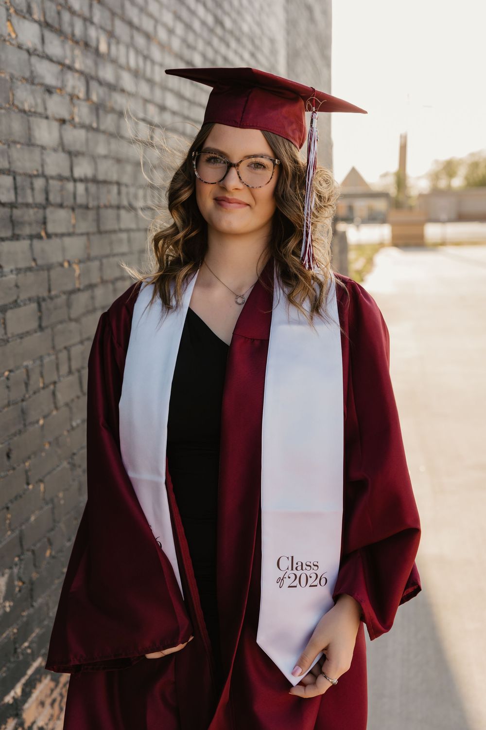 A girl wearing cap and gown and glasses poses for senior portraits in front of black brick in downtown Tulia, Texas.