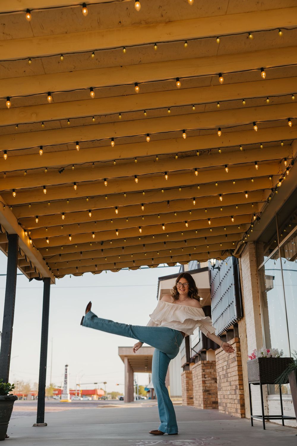 A fun senior portrait featuring a girl kicking her leg and laughing in downtown Tulia, Texas under string lights.