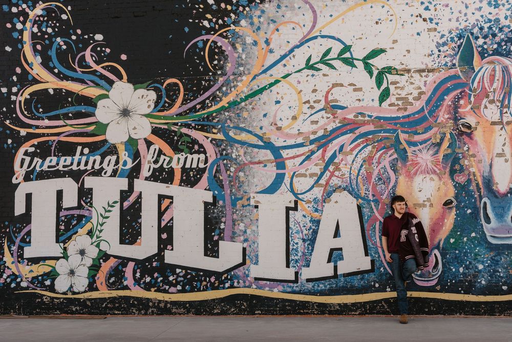 Senior guy wearing letterman jacket poses in front of popular mural in downtown Tulia, Texas for his senior photos.