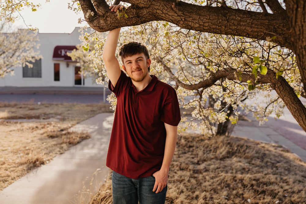 A guy wearing red shirt poses under tree with white flowers in downtown Tulia, Texas for his senior photos.