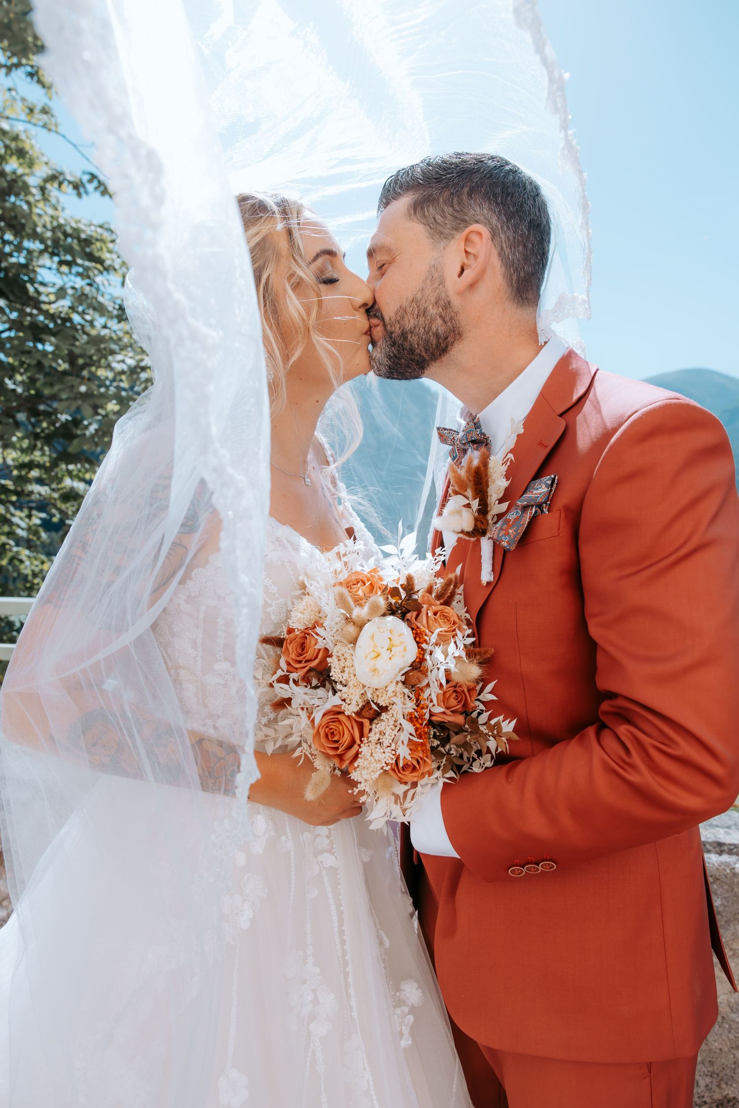 Bride and groom sharing a romantic kiss under a flowing veil, holding an orange and white bouquet outdoors.