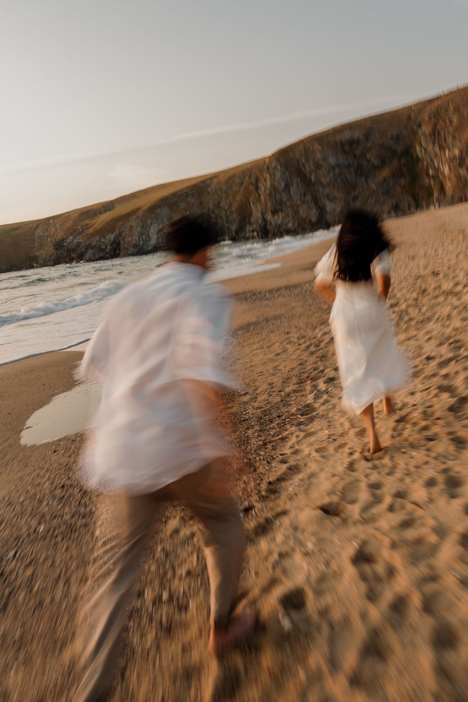 Blurred couple in white outfits running on a sandy beach at sunset with rocky cliffs in the background.