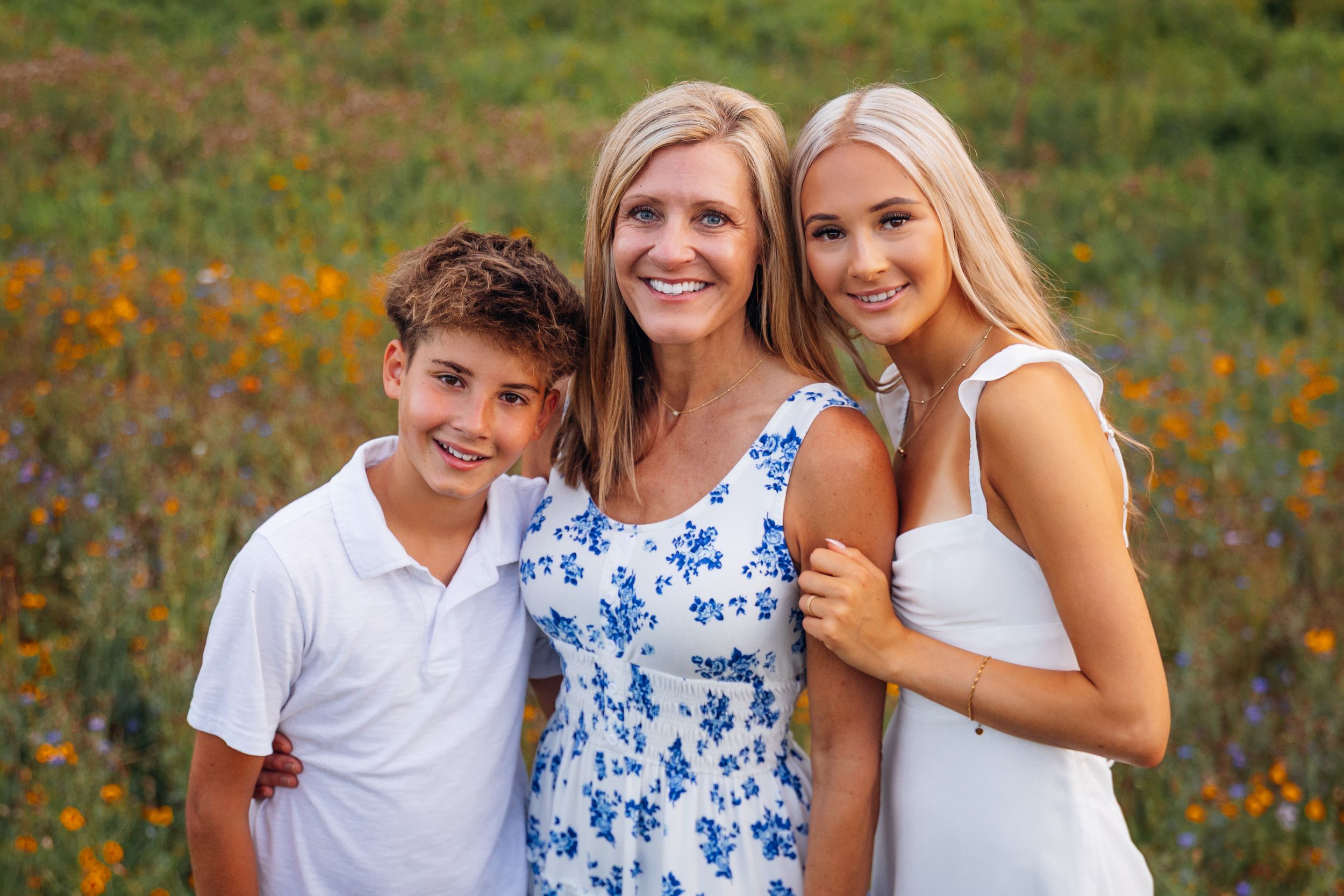 A smiling mother poses with her teenage daughter and young son in a sunny wildflower field.