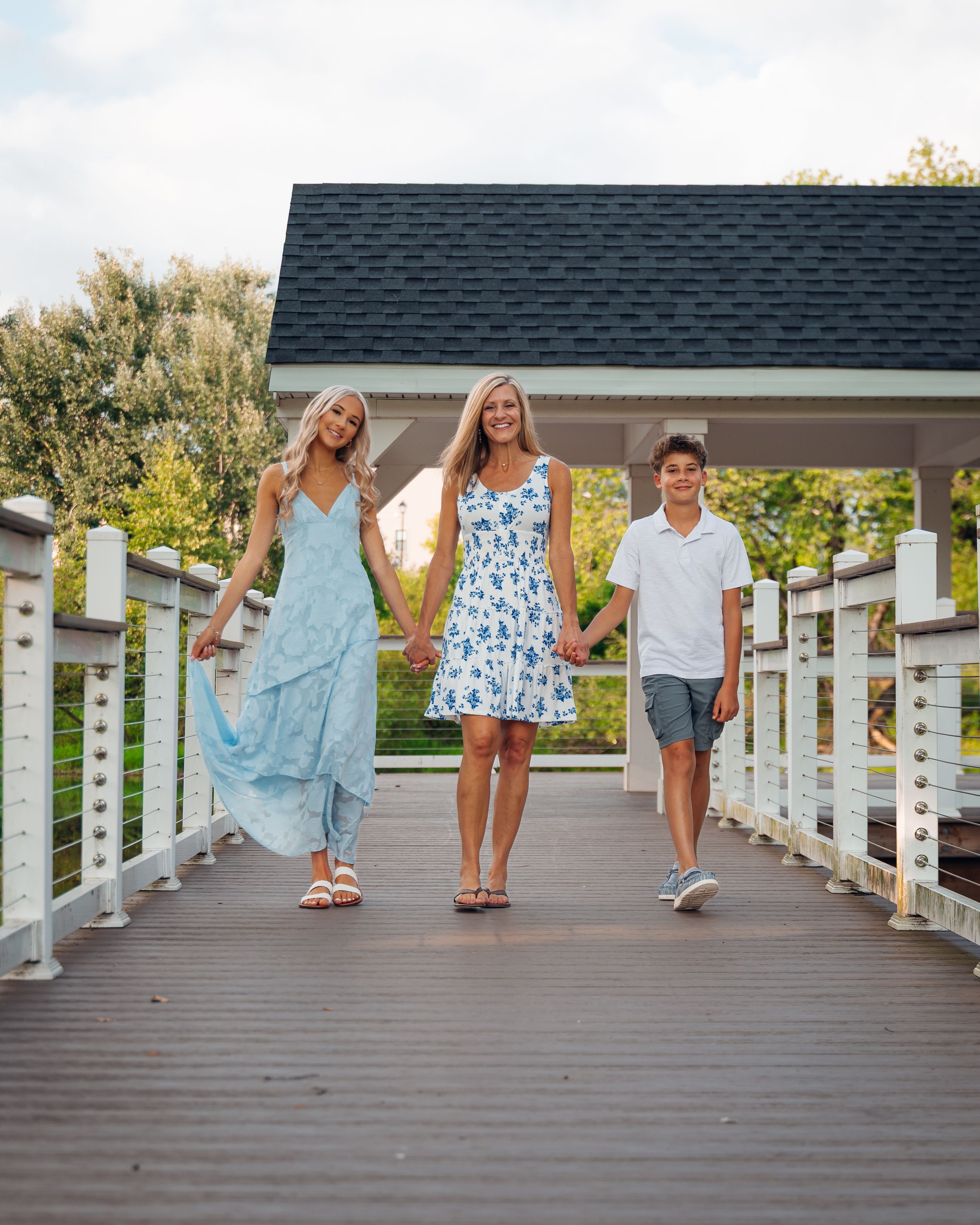 Three people walking hand-in-hand on a wooden bridge, wearing blue outfits, with a gazebo and trees in the background.