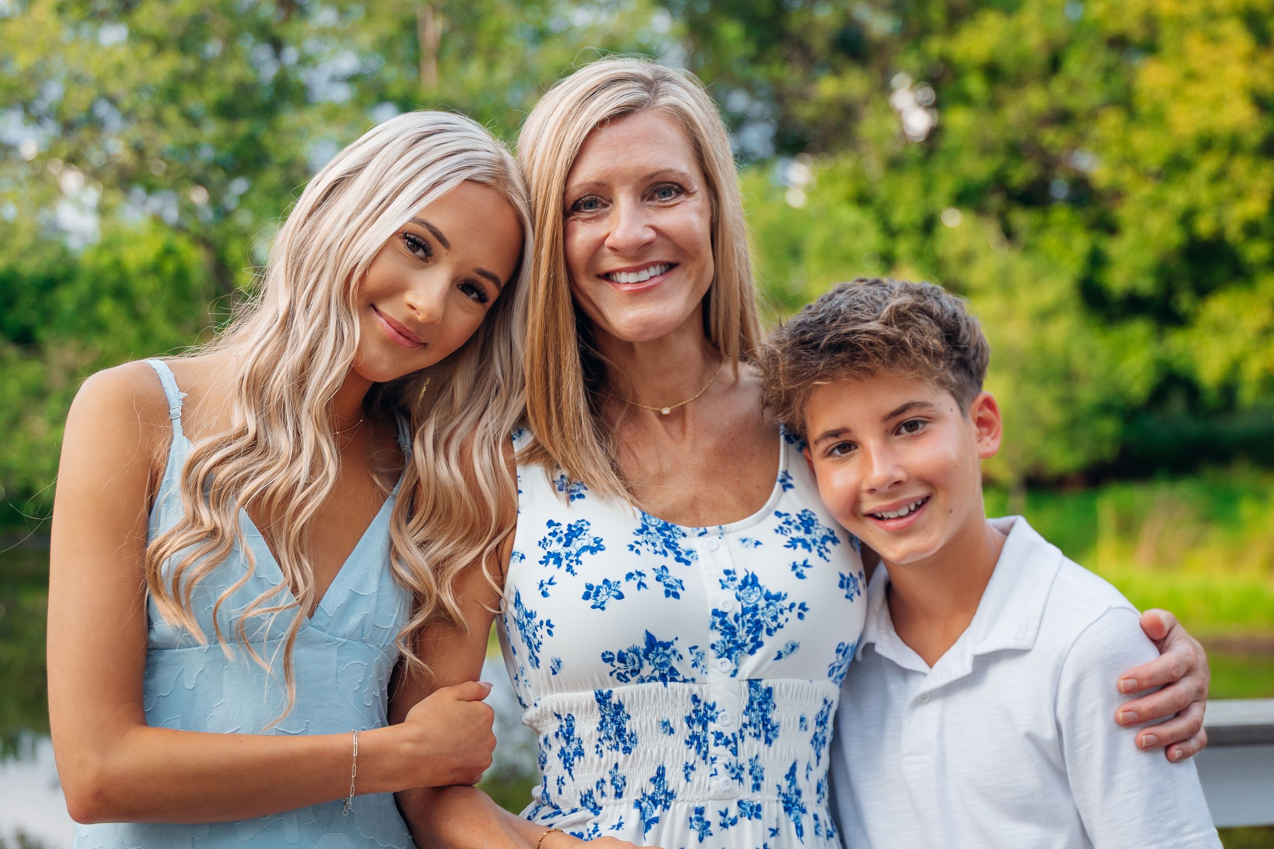 A smiling woman with two children, a teenage girl in a blue dress and a young boy, posing outdoors.