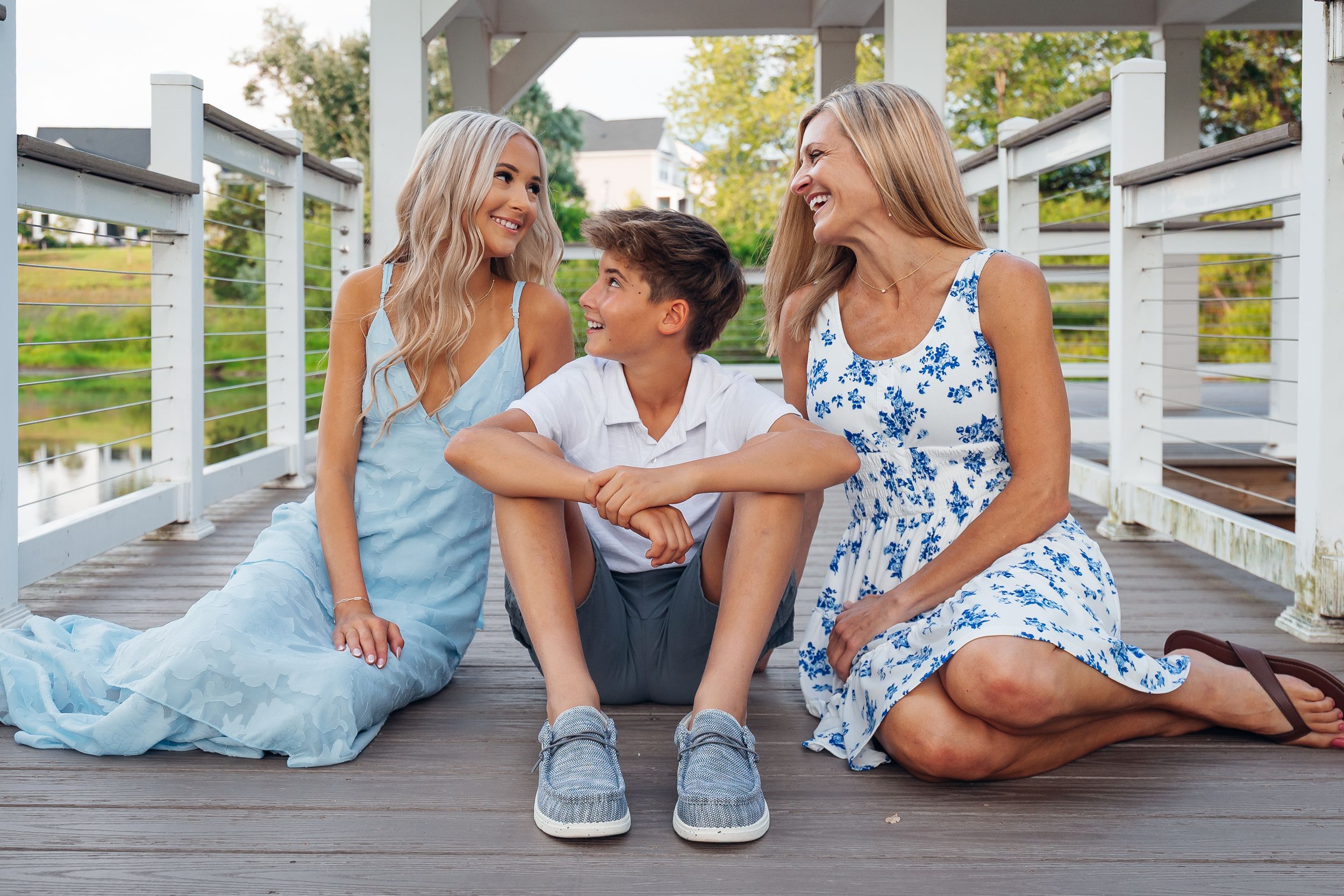 Two women and a boy sit laughing together on a wooden deck, wearing light blue and white outfits outdoors.