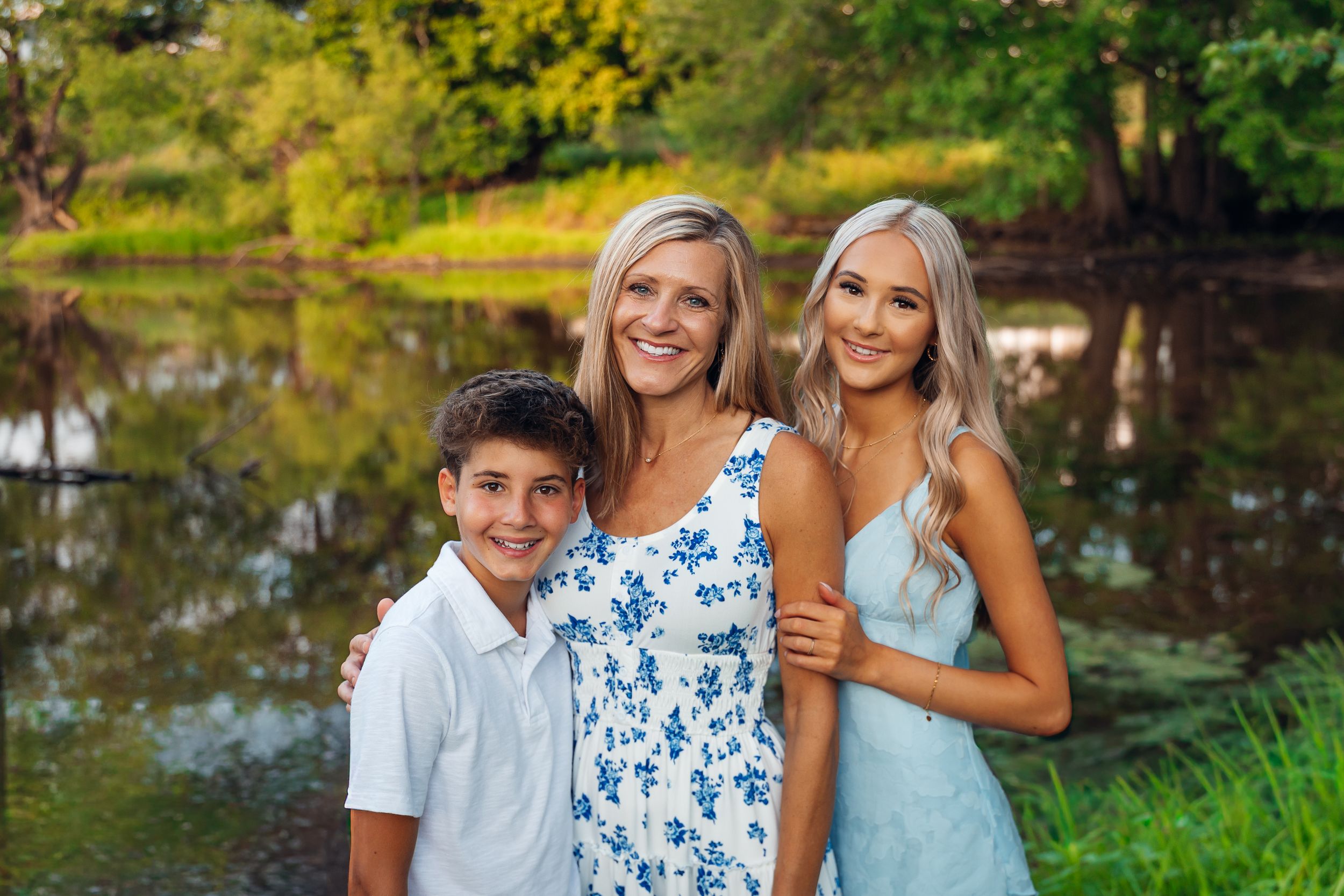 A smiling woman in a floral dress poses outdoors with two children near a pond surrounded by lush greenery.