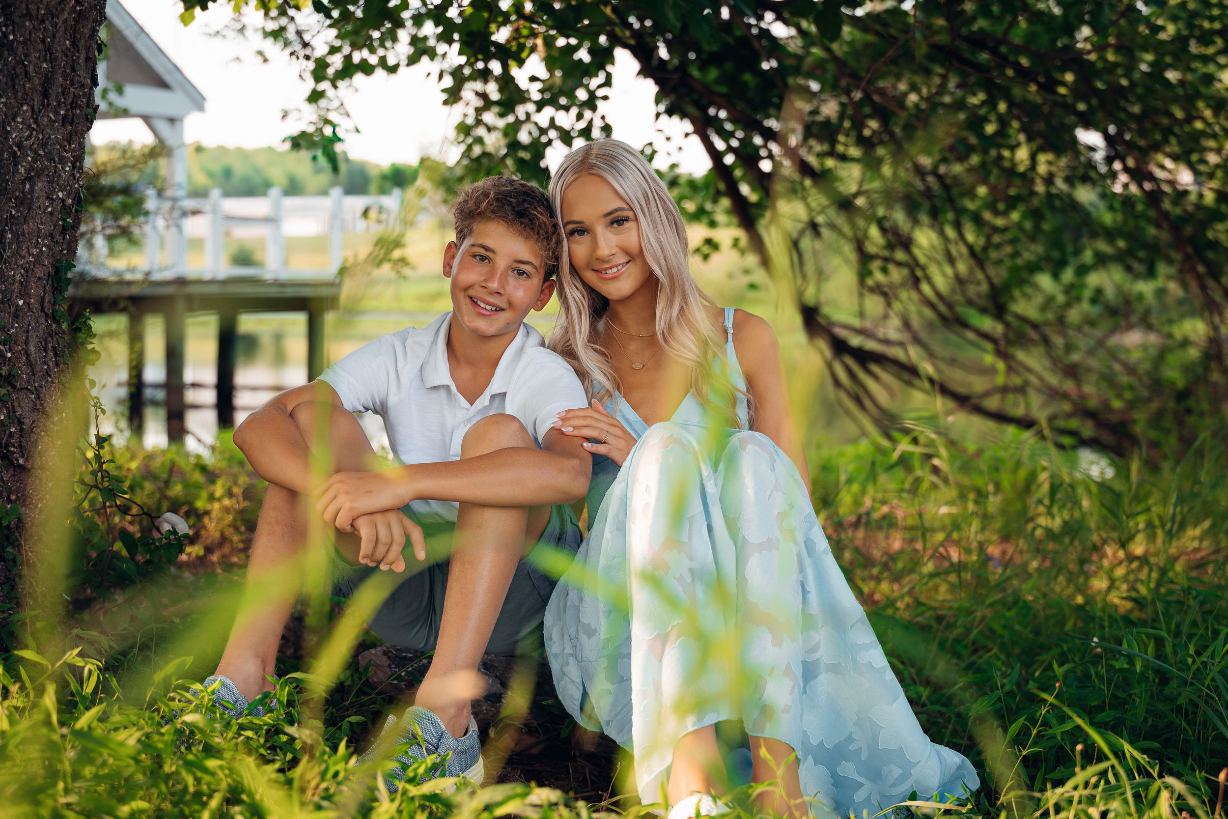A young boy and blonde girl in a blue dress pose together outdoors under a tree in a lush garden setting.