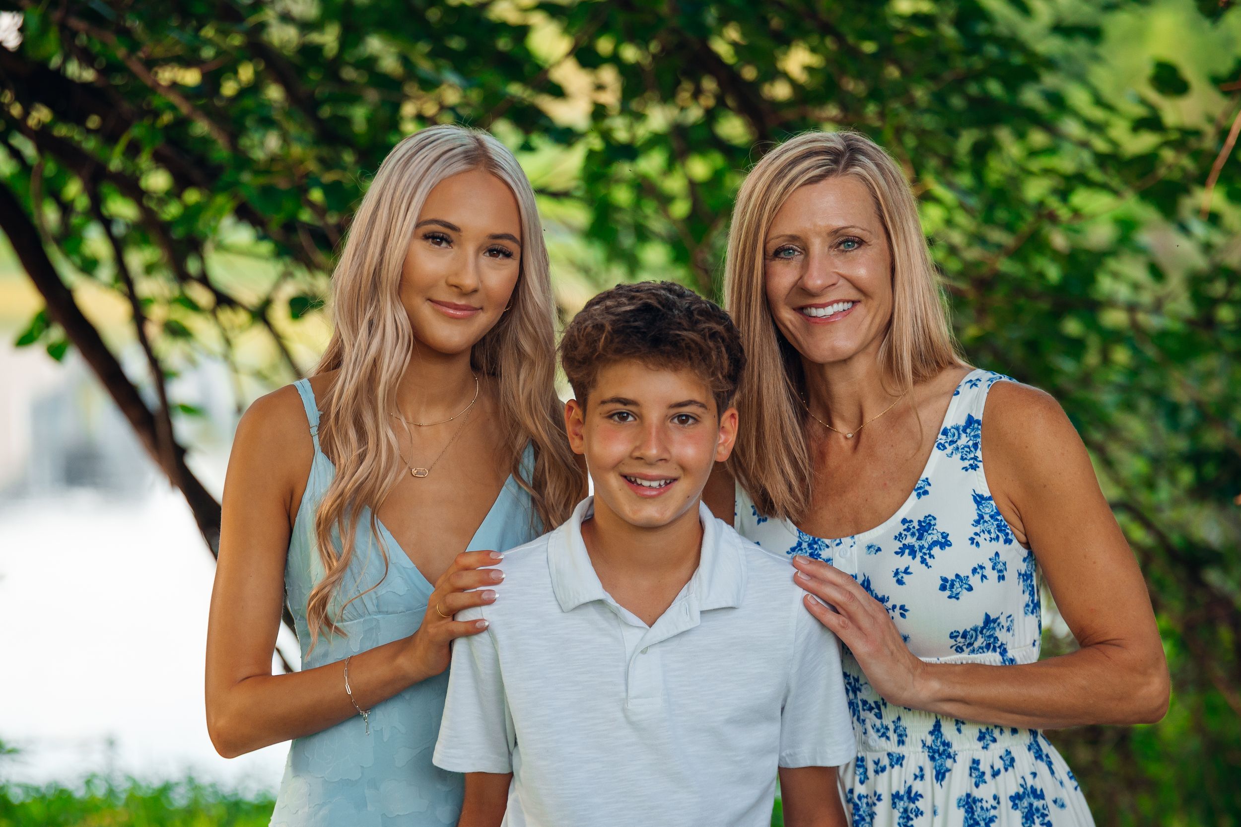 A smiling woman and teenage girl in blue outfits pose with a young boy outdoors under green trees.