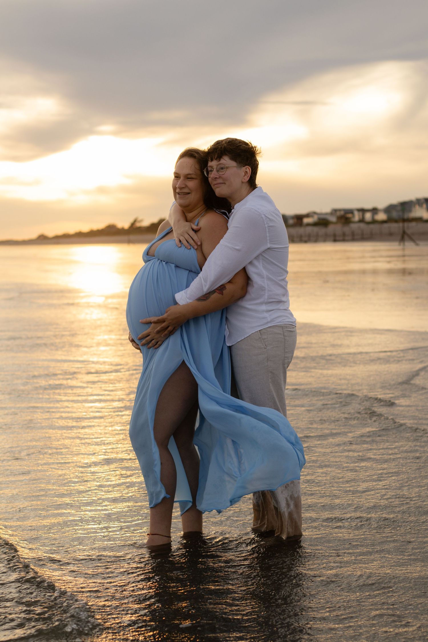 Two women embrace on a beach at sunset, one pregnant in a flowing blue dress, standing in shallow water.