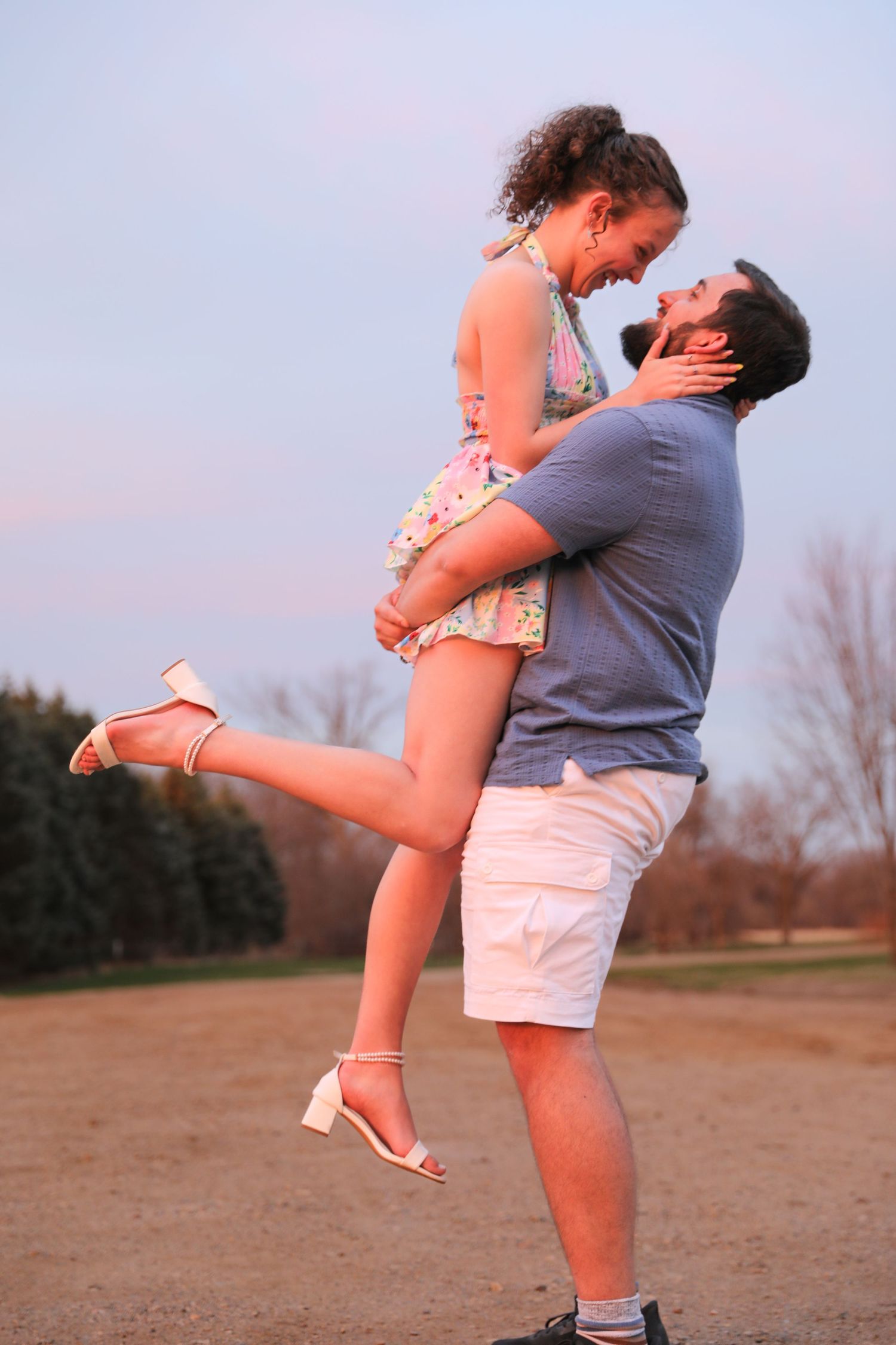A man joyfully lifts a smiling woman in a floral dress outdoors at sunset in a romantic couple moment.