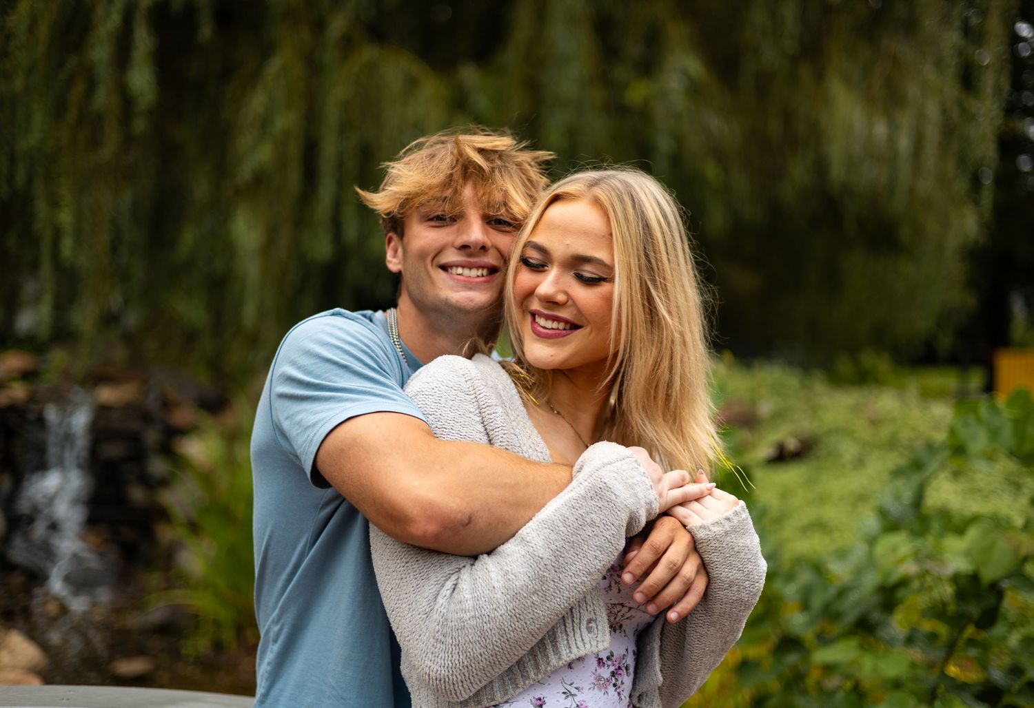 Happy young couple hugging and laughing outdoors near a waterfall and willow tree.