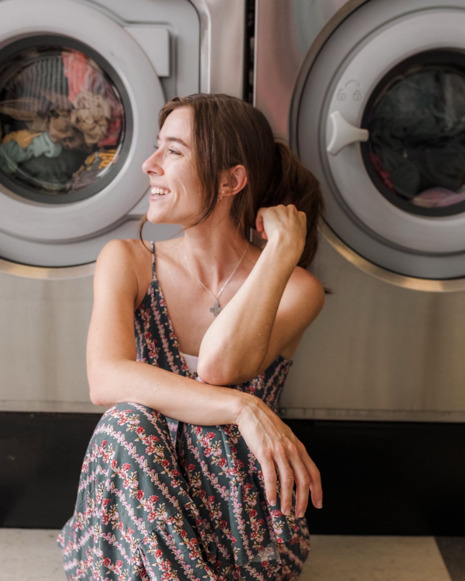 Smiling woman in floral dress sitting on floor at laundromat with washing machines in background.