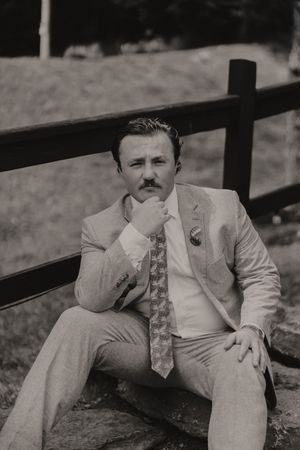 Black and white portrait of groom with mustache in light suit sitting by wooden fence outdoors.