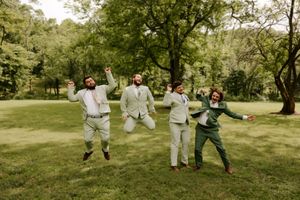 Four groomsmen jumping and posing joyfully outdoors in light suits on green lawn surrounded by trees.