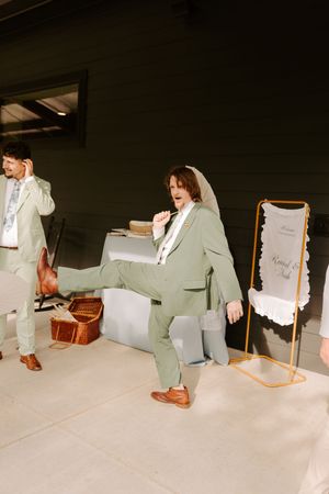 Groomsman in sage green suit kicks leg up playfully at wedding reception near welcome sign.