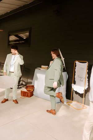 Two groomsmen in sage green suits and brown dress shoes prepare for a wedding reception near a welcome sign.