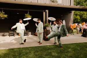 Groomsmen in sage green suits dance playfully with parasols on a patio during an outdoor wedding celebration.