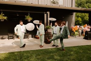 Groomsmen in sage green suits playfully pose with parasols on a garden patio during a wedding celebration.