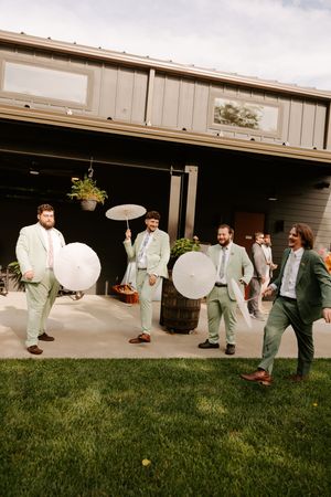 Groomsmen in mint green suits holding white parasols laugh outside a rustic barn wedding venue.