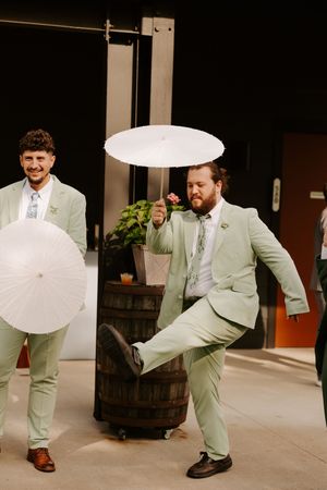 Groomsmen in matching sage green suits kick legs up while holding white parasols at wedding.