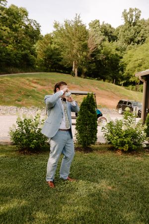 Groom in light blue suit uses instant camera to photograph outdoors near garden shrubs.