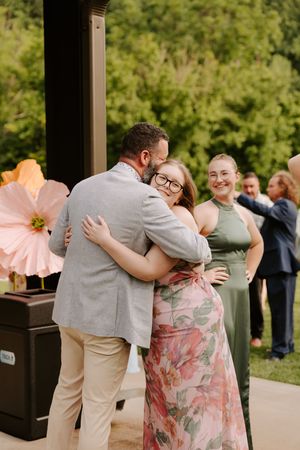 A man in a gray blazer shares a warm hug with a woman in a floral dress at an outdoor wedding celebration.