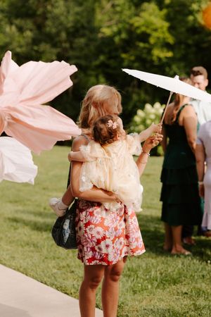 Woman holding child with pink fabric and white parasol at outdoor summer event.