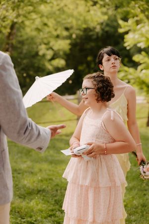 Girl in pink dress holds white parasol outdoors while another woman in yellow dress stands behind her.