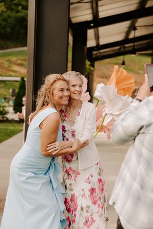Two women in elegant dresses smile and pose together at an outdoor wedding event with large decorative flowers.