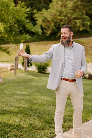 A bearded man in a light gray blazer smiles while holding a drink and a card at an outdoor wedding.