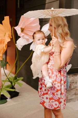 Woman in floral dress holding baby under white parasol near large decorative flowers outdoors.