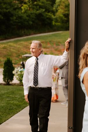 Older man in white dress shirt and tie leaning against a wall at an outdoor wedding reception.