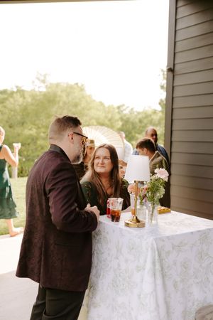 Guests mingle at an outdoor wedding reception cocktail hour near a decorated table with a lamp and fresh flowers.