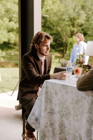 Young man in brown suit sitting at floral tablecloth table outdoors, gazing thoughtfully at a garden event.
